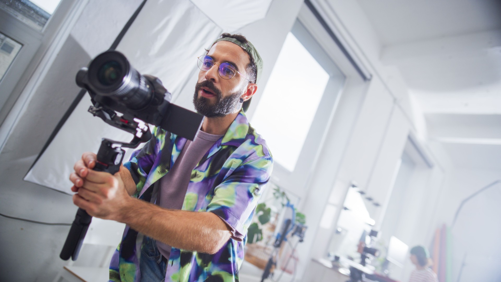 Focused Young Latin Male Videographer With Beard, Filming In A Bright Studio, Holding A Professional Camera, Surrounded By Modern Filmmaking Equipment, Engaged In A Creative Production Process.